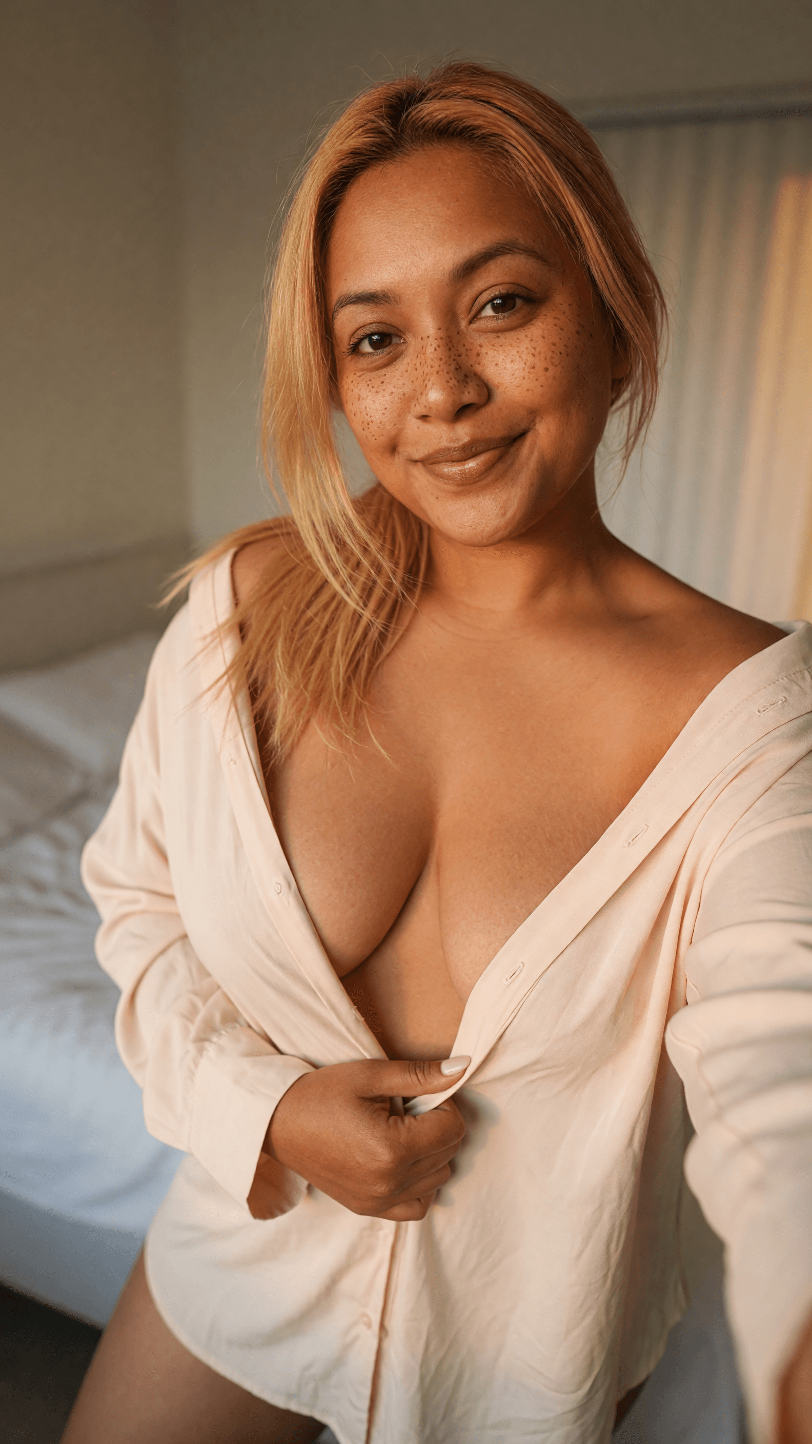 Blonde unbuttons pink shirt on bed