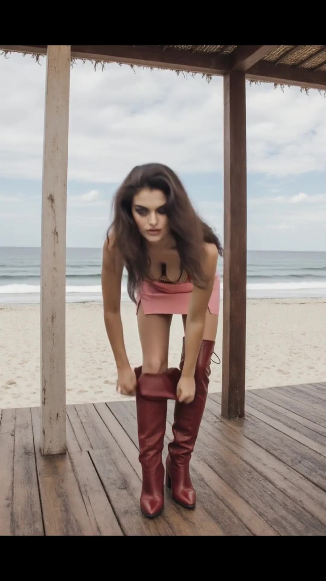 Blonde in pink mini skirt pulls on red boots beach deck